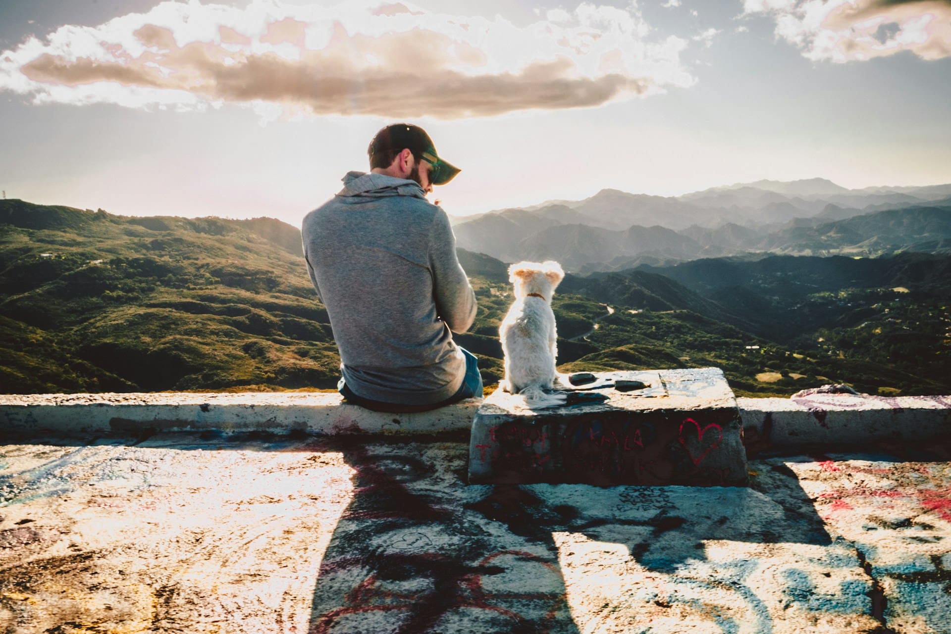 man wearing hat sitting with dog on ledge overlooking mountains